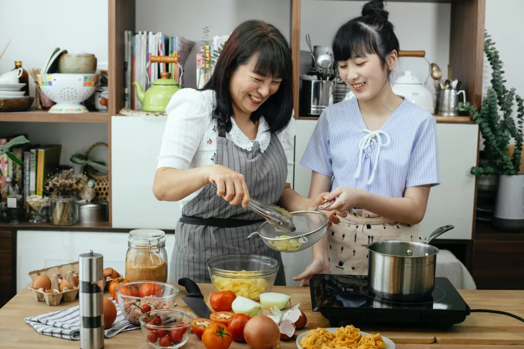 Laughing Asian mother and daughter preparing pasta in kitchen